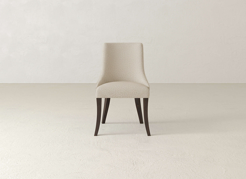 The James+James Willow Dining Chair, armless and upholstered in light beige fabric with dark wooden legs, sits centered in a minimalist, neutral-toned room with a textured floor and plain walls.