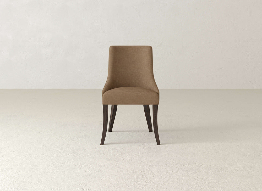 A brown James+James Willow Dining Chair with dark wooden legs sits in the center of a plain, light-colored room featuring a textured floor and off-white walls.
