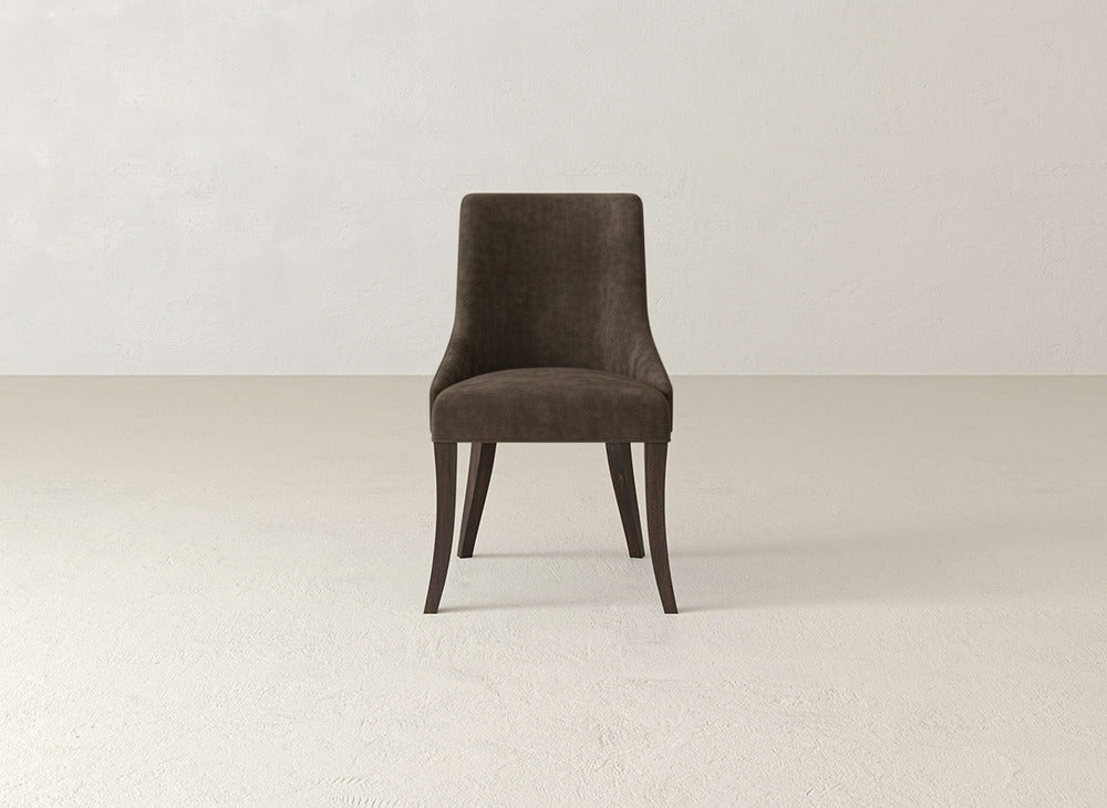 Centered against a plain, light-colored wall and floor, a modern James+James Willow Dining Chair features dark brown upholstery and wooden legs.