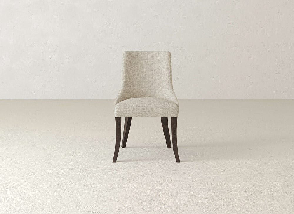A James+James Willow Dining Chair with light beige upholstery and dark wooden legs sits centered in a neutral, empty room with textured walls and floor.