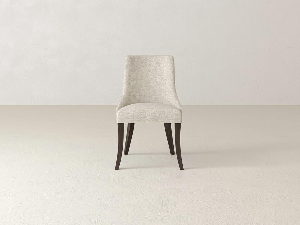 A beige upholstered chair with dark wooden legs stands in a minimalist, light-colored room. The chair has a smooth, curving backrest and a simple, elegant design.