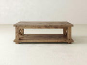 The Carter X Coffee Table by James+James is a rustic wooden piece with a rectangular shape and a lower shelf for additional storage. It features a harvest wheat finish and sturdy legs, reflecting a simple, minimalist design. The background includes a plain, light-colored wall and floor.