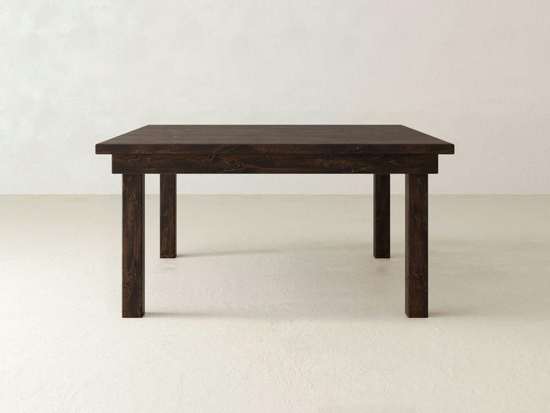 A Farmhouse Square Dining Table from James+James is placed against a plain light-colored background, showcasing its sturdy and minimalist design. The solid hardwood surface and legs display a natural wood grain texture, adding a touch of rustic elegance.