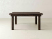 A Farmhouse Square Dining Table from James+James is placed against a plain light-colored background, showcasing its sturdy and minimalist design. The solid hardwood surface and legs display a natural wood grain texture, adding a touch of rustic elegance.