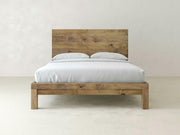 The James+James Sawyer Bed in Harvest Wheat on Alder features a minimalist wooden frame and matching headboard, dressed in white bedding and pillows, set against a plain light-colored wall and floor.