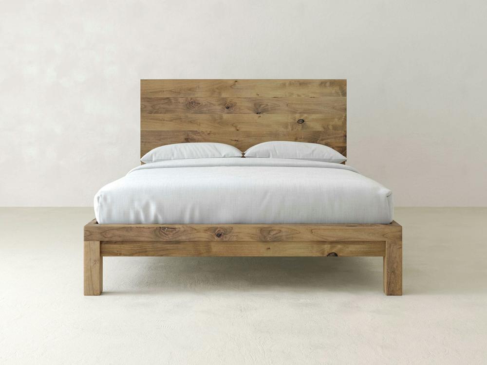 The James+James Sawyer Bed in Harvest Wheat on Alder features a minimalist wooden frame and matching headboard, dressed in white bedding and pillows, set against a plain light-colored wall and floor.