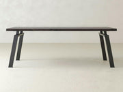 The James+James Fulton Desk, a minimalist wooden table featuring a dark top and angled black metal legs, is showcased in a plain, light-colored room with a concrete floor and textured walls. The design exudes industrial elegance with its sleek lines and sturdy appearance. Customization options are available.