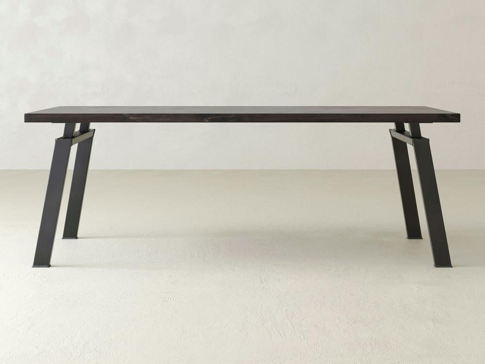 The James+James Fulton Desk, a minimalist wooden table featuring a dark top and angled black metal legs, is showcased in a plain, light-colored room with a concrete floor and textured walls. The design exudes industrial elegance with its sleek lines and sturdy appearance. Customization options are available.