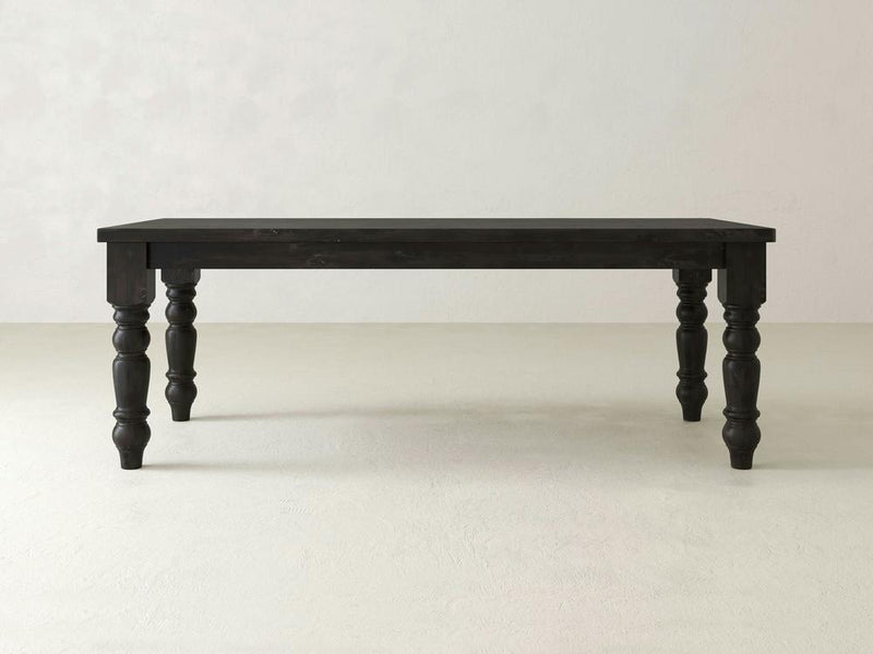 A James+James Baluster Dining Table in the Charred Ember finish, featuring four ornately carved legs, stands on a light-colored floor against a plain, light-colored wall. The table has a rectangular shape and a smooth surface. The overall design is classic and elegant.