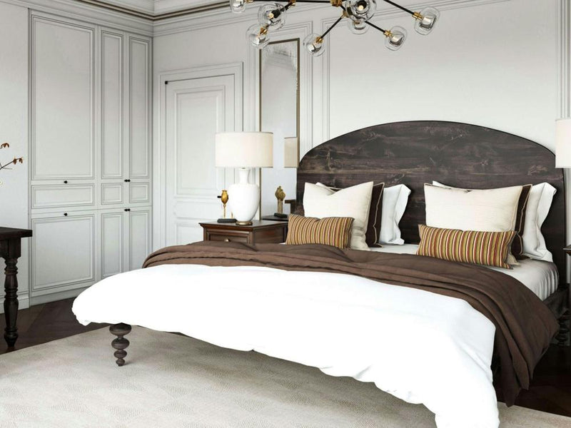 A cozy bedroom with the James+James Olivia Arch Bed in Charred Ember, adorned with white pillows and brown striped cushions. The space has a white paneled wall, ornate chandelier, and wooden furniture, offering a warm and elegant atmosphere.