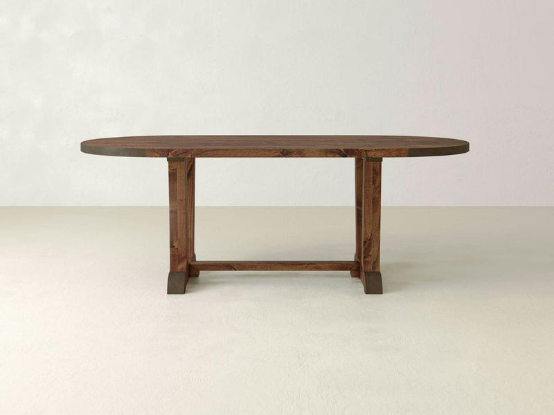 The Thaden Oval Dining Table by James+James boasts a rich, dark brown finish. With its simple, clean lines and modern design, this rectangular wooden table features sturdy legs connected by a support beam near the base. Crafted from rustic knotty alder hardwood, its smooth surface beautifully displays the natural wood grain against a plain, light-colored wall.