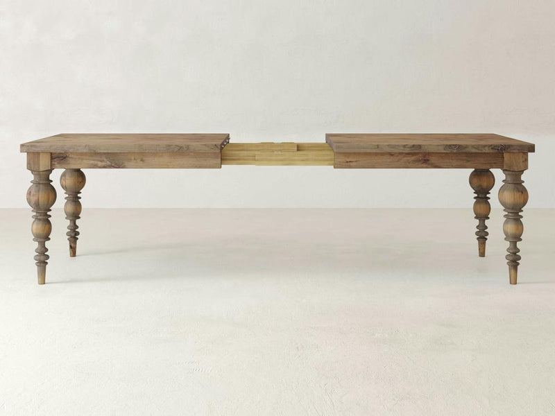 The Bailey Expandable Dining Table - Harvest Wheat by James+James is displayed with its ornate, turned legs and a central extension mechanism, ready to accommodate an additional leaf. The table features a rustic finish and stands on a plain, light-colored floor against a neutral background.