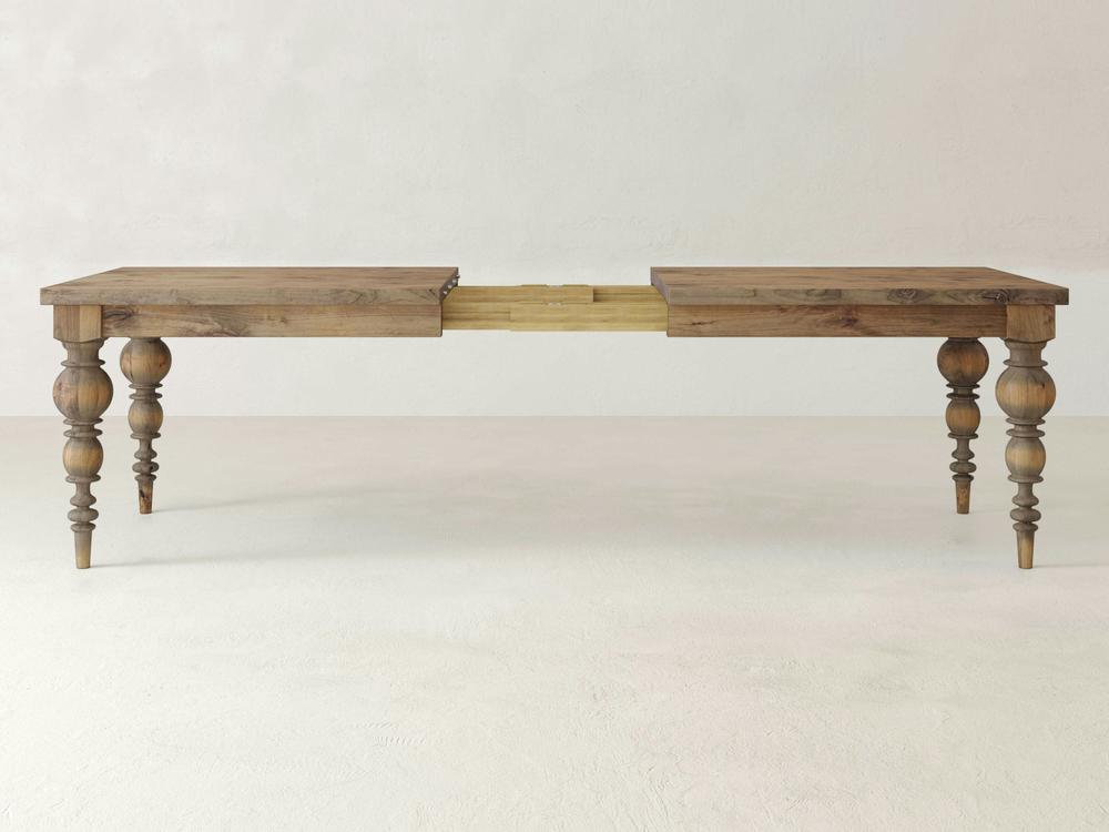 The Bailey Expandable Dining Table - Harvest Wheat by James+James is displayed with its ornate, turned legs and a central extension mechanism, ready to accommodate an additional leaf. The table features a rustic finish and stands on a plain, light-colored floor against a neutral background.
