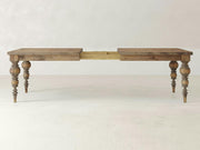 The Bailey Expandable Dining Table - Harvest Wheat by James+James is displayed with its ornate, turned legs and a central extension mechanism, ready to accommodate an additional leaf. The table features a rustic finish and stands on a plain, light-colored floor against a neutral background.