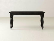 A James+James Abigail Sofa Table - Charred Ember with four ornate, carved legs stands alone on a light-colored floor against a bare, off-white wall. The tabletop has a simple rectangular shape, providing a contrast with the detailed craftsmanship of the legs.