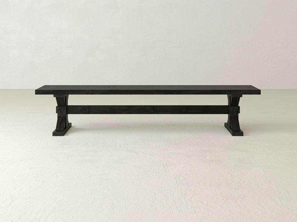 The James+James Trestle Bench in Charred Ember on Alder, featuring a simple and sturdy design, sits on a light floor against a plain white wall.