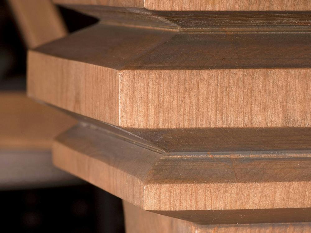 Close-up of the Heirloom Expandable Dining Table - Harvest Wheat by James+James, highlighting its geometric, wooden architectural detail featuring angular, protruding edges with precise symmetrical cuts. The wood grain is clearly visible, showcasing its natural texture and warm, reddish-brown hue against a blurred background.