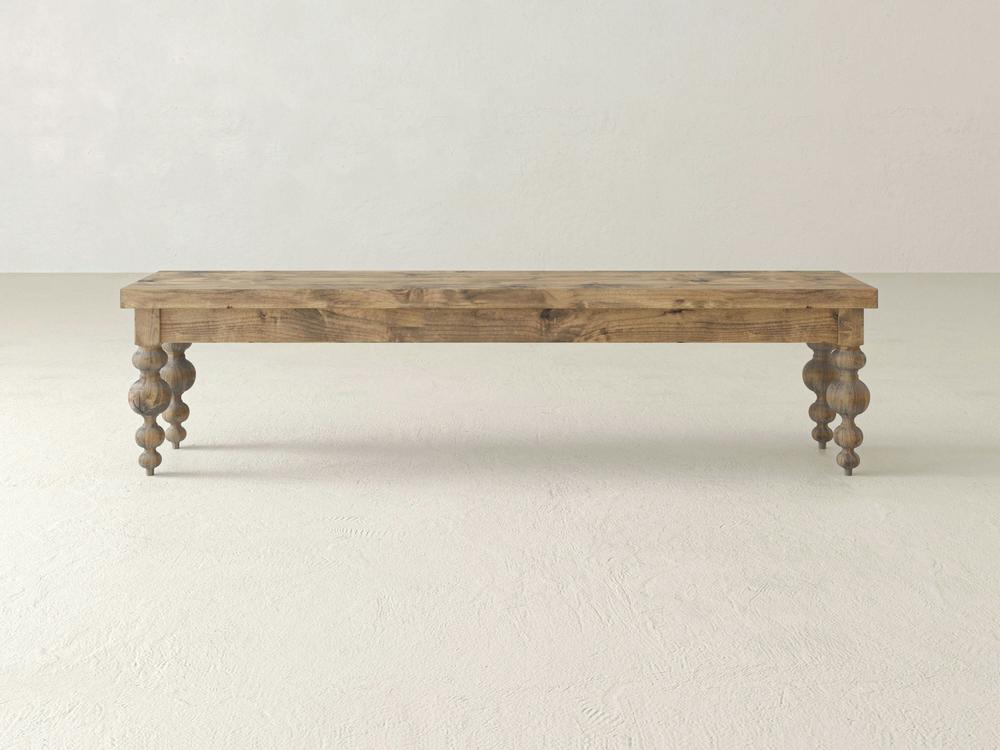 The Olivia Bench - Harvest Wheat on Alder by James+James features a rectangular wooden top and four ornately carved legs, displayed against a plain light-colored wall and floor.