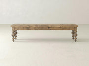 The Olivia Bench - Harvest Wheat on Alder by James+James features a rectangular wooden top and four ornately carved legs, displayed against a plain light-colored wall and floor.