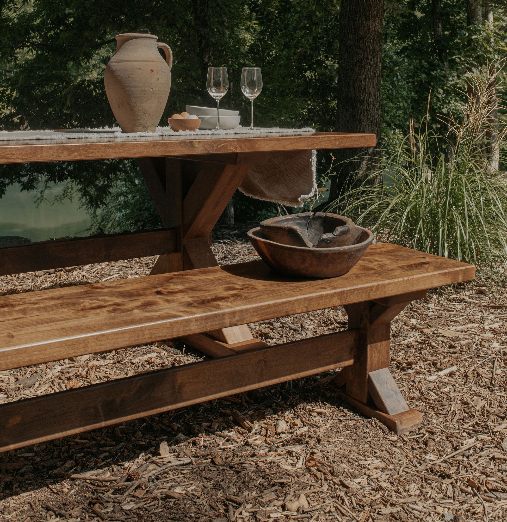A James+James Trestle Bench - Tobacco on Alder sits outdoors amid trees, topped with a ceramic pitcher, wine glasses, a bowl of bread, and lace runner; a wooden bowl with water rests nearby amidst lush greenery.