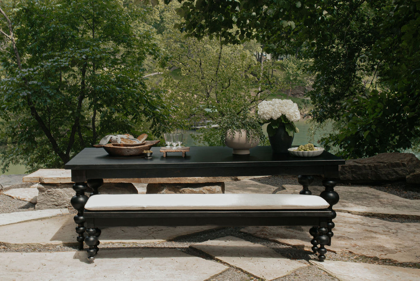 The Olivia Dining Table in Harvest Wheat by James+James sits on a stone patio surrounded by lush trees, elegantly set with flowers, drinks, food, and grapes atop its surface, overlooking a peaceful natural landscape.