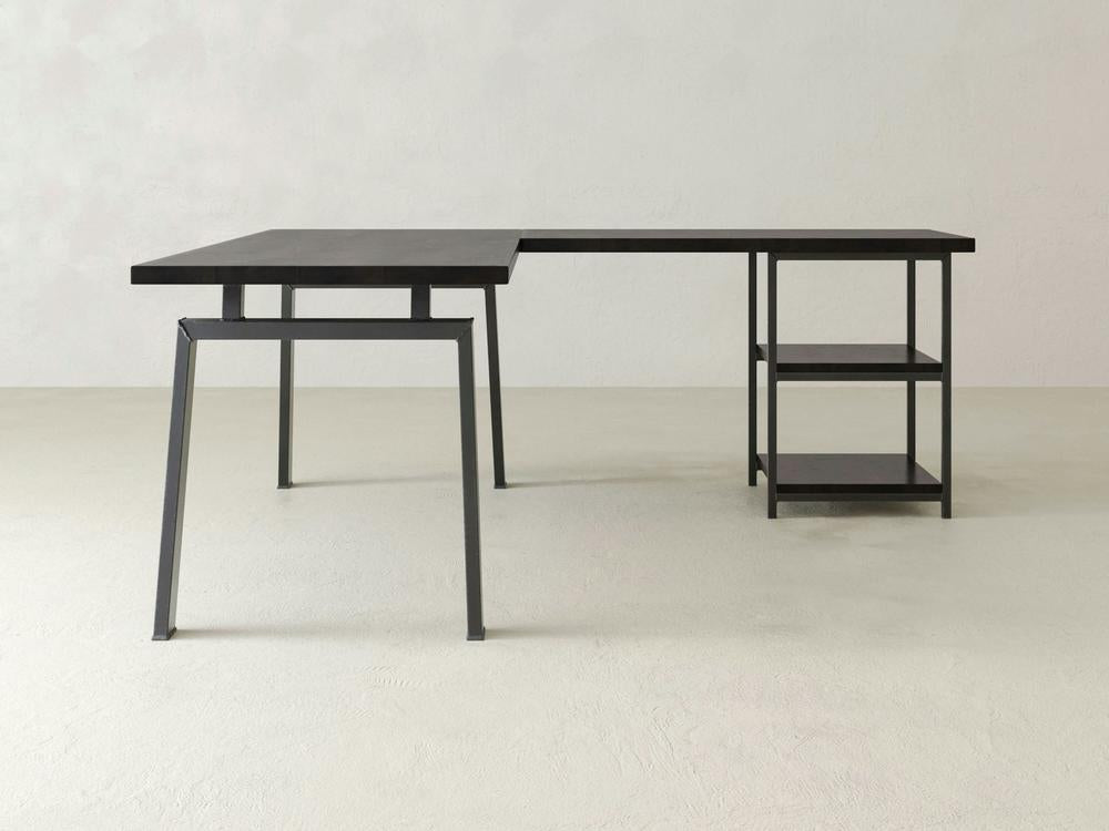 The James & James Fulton Corner Desk - Charred Ember is a minimalist L-shaped desk with sleek metal legs and dark wood surfaces, featuring two open shelves on one side set against a plain, light background.