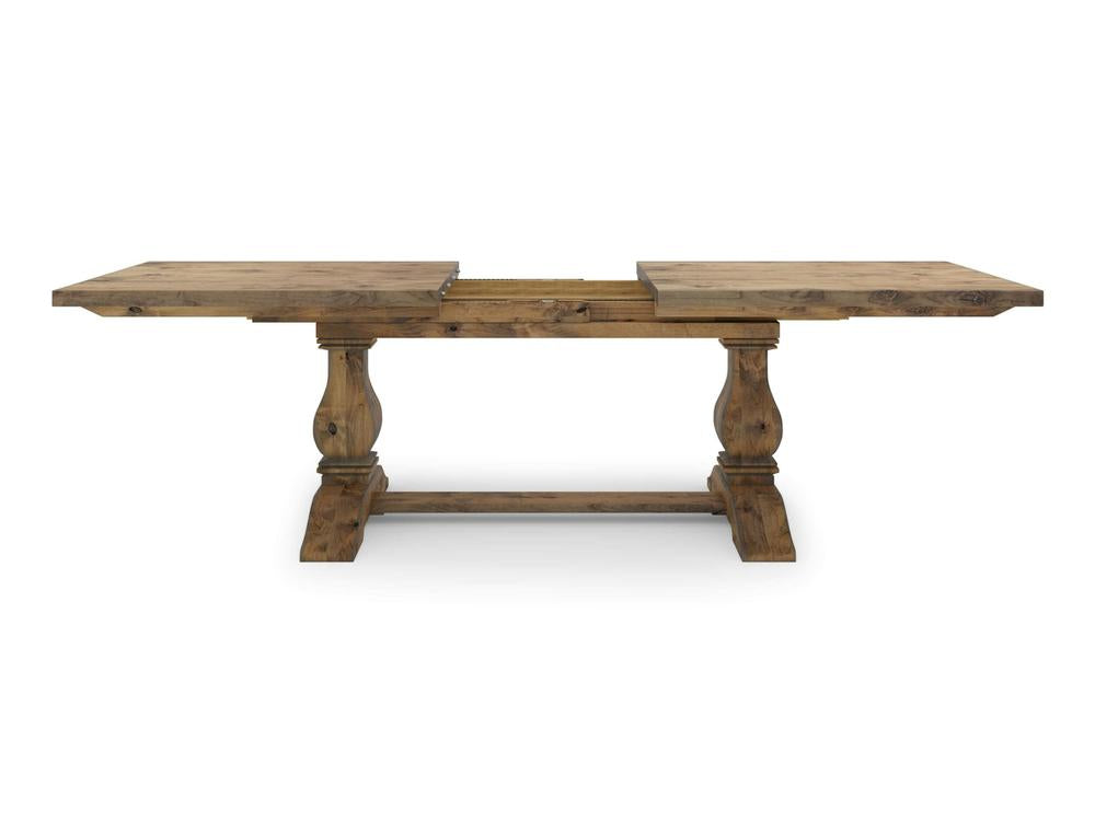 The Heirloom Expandable Dining Table - Harvest Wheat by James+James is a rustic wooden dining table with two extended leaves. It features a sturdy, trestle-style base supported by intricately carved legs. The wood's weathered, natural finish gives it a vintage and farmhouse aesthetic.