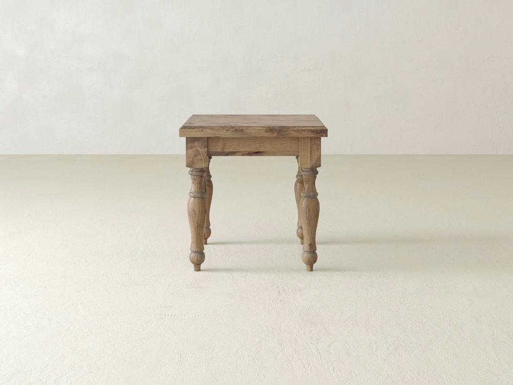 An Abigail End Table - Harvest Wheat by James+James with a natural finish and a rustic design is centered in the image. The table features four carved legs and a simple rectangular top. The background showcases a minimalistic, off-white room with smooth walls and floor.