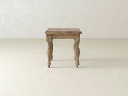 An Abigail End Table - Harvest Wheat by James+James with a natural finish and a rustic design is centered in the image. The table features four carved legs and a simple rectangular top. The background showcases a minimalistic, off-white room with smooth walls and floor.