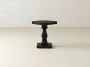 The Vivien Side Table - Charred Ember by James+James features a dark wooden circular top resting on an ornately carved, sturdy single leg, all supported by a square base. The table is showcased against a plain, light-colored background.