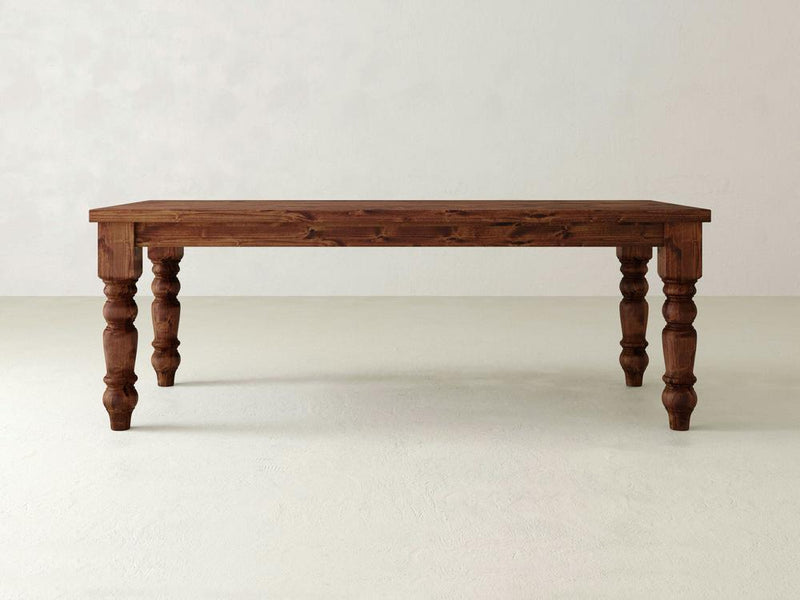 The Baluster Dining Table - Tuscany by James+James features a rectangular wooden top and four ornately carved, turned legs. It boasts a natural wood finish and is showcased against a plain, light-grey wall on a light-colored floor.