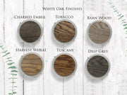 Image showing six wood finishes on a white background: Charred Ember, Tobacco, Barn Wood, Harvest Wheat, Tuscany, and Deep Grey. Each sample is circular, framed in metallic rings, with green foliage on the sides.