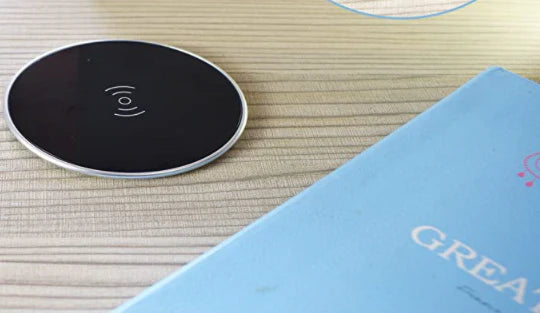 The James+James Standard Desk Add-On, a black wireless charging pad with a minimalist design, sits on a wooden surface. To its right lies a partially visible light blue book titled "GREAT" with a circular emblem at the top. The charging pad reflects light elegantly.