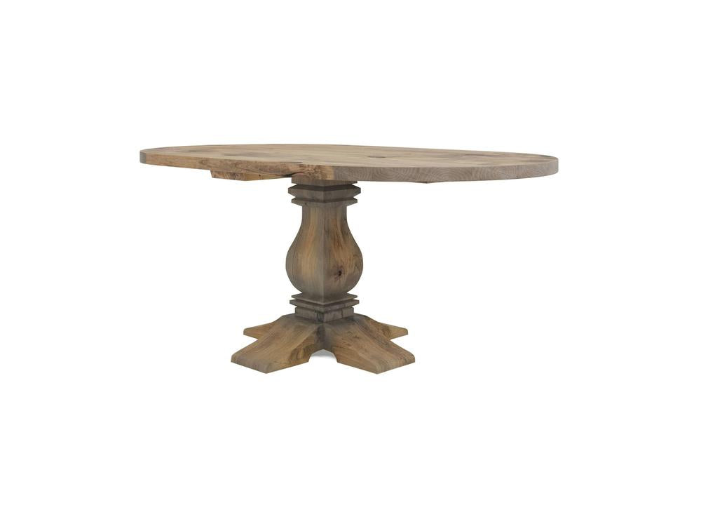 Heirloom Oval Dining Table - Harvest Wheat by James+James. This rustic wooden table boasts an oval top and a single, elegantly carved pedestal base. The wood showcases a natural, slightly worn finish, enhancing its vintage charm. The table is supported by a sturdy, cross-shaped base.