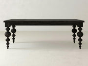 The Olivia Dining Table - Charred Ember by James+James is a dark wooden table with intricately carved legs featuring both spherical and cylindrical shapes. Its rectangular tabletop stands out against a plain, light-colored wall, presenting a minimalist yet elegant design.