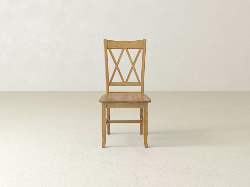A single Double X-Back Dining Chair - Harvest Wheat by James+James is centrally placed against a plain, light-colored background. The chair has a rectangular seat and a backrest featuring a double-crossed design. The floor and wall are both light beige.