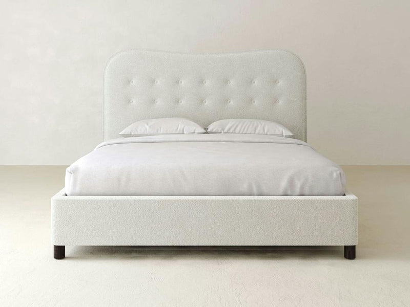 The Florence Bed – Boucle by James & James features a minimalist white design with a tufted headboard and two plush pillows, upholstered in textured boucle. It stands on solid maple feet against a plain wall, crafting a serene, clean atmosphere.