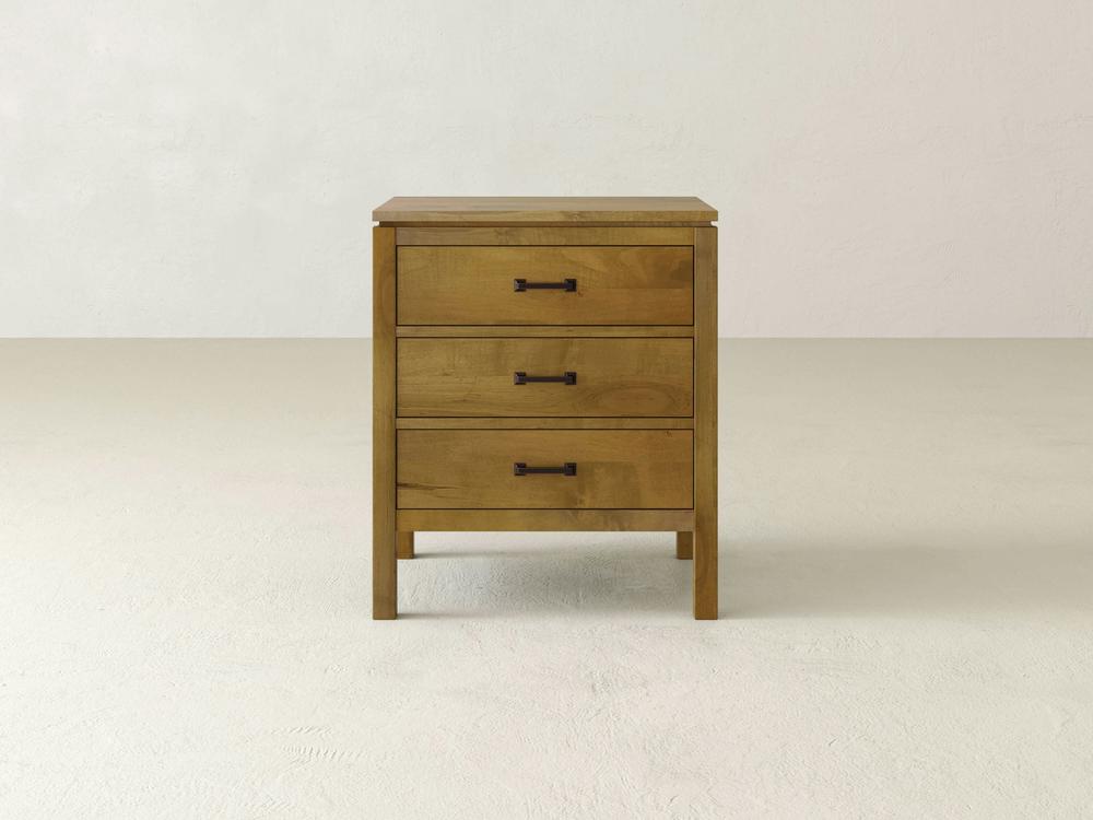 The James & James Jensen 3 Drawer Nightstand is crafted from solid maple hardwood with a light brown finish. It stands elegantly on a plain floor against a white wall, offering ample bedside storage with three drawers, each featuring simple black metal handles.