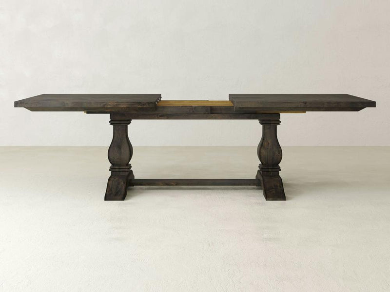 The Heirloom Expandable Dining Table by James+James, showcased with its leaves extended and finished in a dark hue, exemplifies rustic elegance. This wooden table features two rectangular support legs connected by a horizontal stretcher and boasts a visible central support section, all set against a plain light-colored wall.