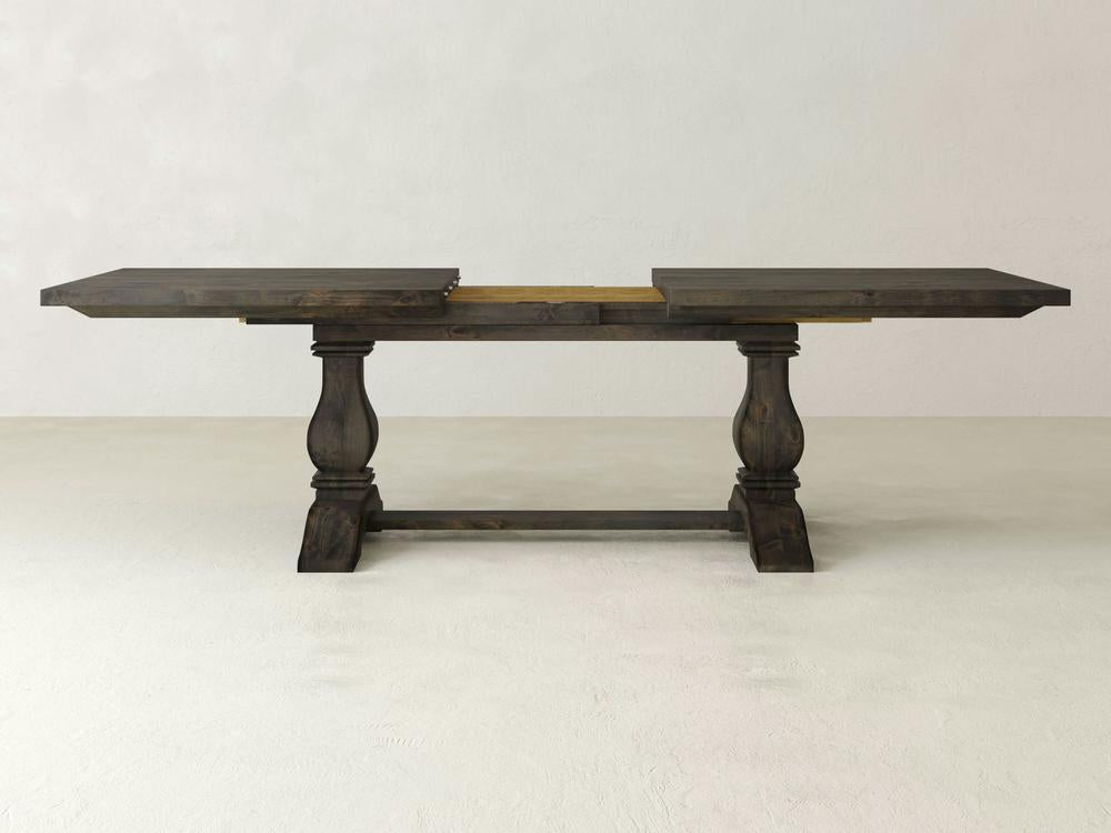 The Heirloom Expandable Dining Table by James+James, showcased with its leaves extended and finished in a dark hue, exemplifies rustic elegance. This wooden table features two rectangular support legs connected by a horizontal stretcher and boasts a visible central support section, all set against a plain light-colored wall.
