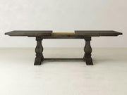 The Heirloom Expandable Dining Table by James+James, showcased with its leaves extended and finished in a dark hue, exemplifies rustic elegance. This wooden table features two rectangular support legs connected by a horizontal stretcher and boasts a visible central support section, all set against a plain light-colored wall.