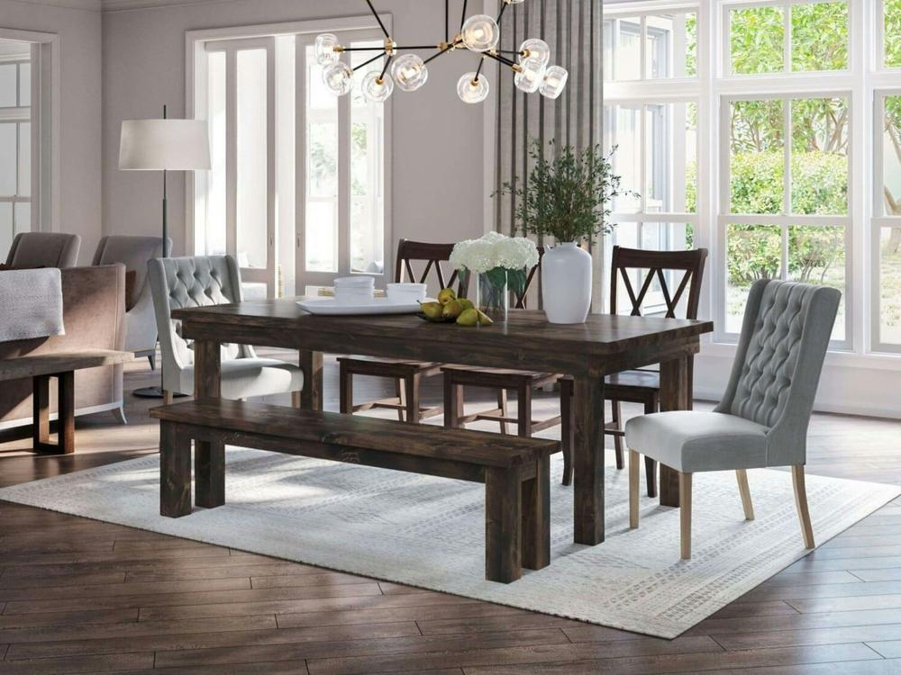 A modern dining room features the James+James Farmhouse Bench—Tobacco on Alder—with a matching dark wood table and chairs on a white rug. Large windows brighten the space, enhanced by flowers, books, fruit, and a modern chandelier above.