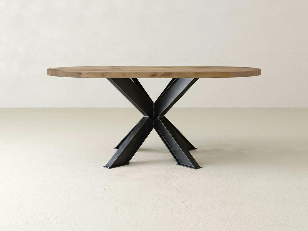 The Shiloh Oval Dining Table by James+James, boasting meticulous artisanal craftsmanship, presents a round wooden top with a sleek, light finish and a black metal X-shaped base. It is stylishly placed on a plain, light-colored floor against a neutral backdrop to achieve an industrial chic aesthetic.