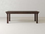 A James+James Farmhouse Dining Table in Tobacco finish, featuring a simple dark wooden design with a rectangular top and four sturdy legs, stands on a light-colored floor against a plain white wall.