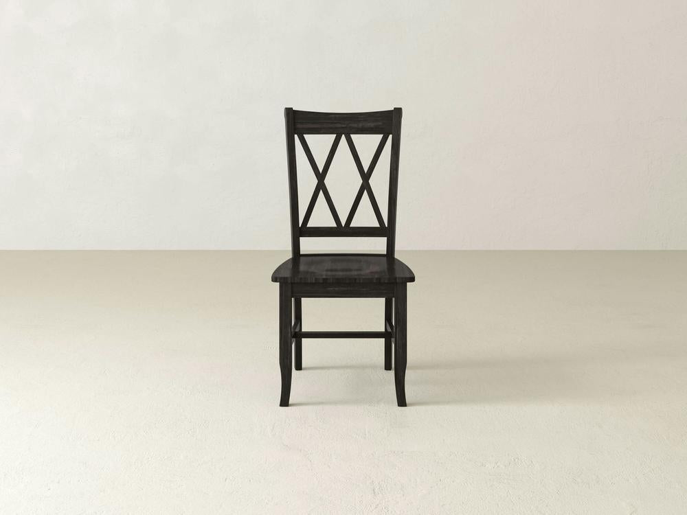 The Double X-Back Dining Chair - Charred Ember by James+James is centered in a minimalistic setting with a light-colored floor and a plain, off-white wall in the background. The chair features a distinctive double X-shaped back design and has a dark, charred ember finish.