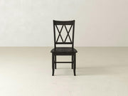 The Double X-Back Dining Chair - Charred Ember by James+James is centered in a minimalistic setting with a light-colored floor and a plain, off-white wall in the background. The chair features a distinctive double X-shaped back design and has a dark, charred ember finish.