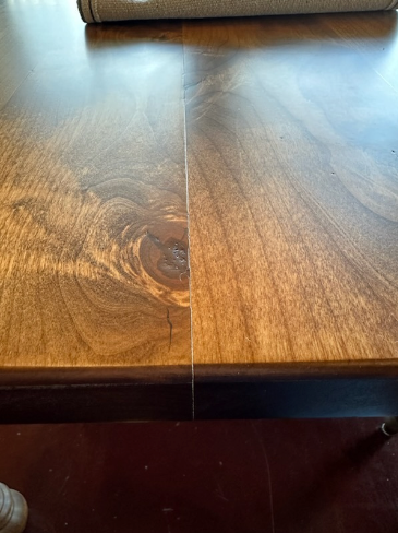 Close-up of a wooden table with a visible split along the grain. The table features a rich brown color with distinct wood patterns and textures. A rolled-up fabric is partially visible on the surface.